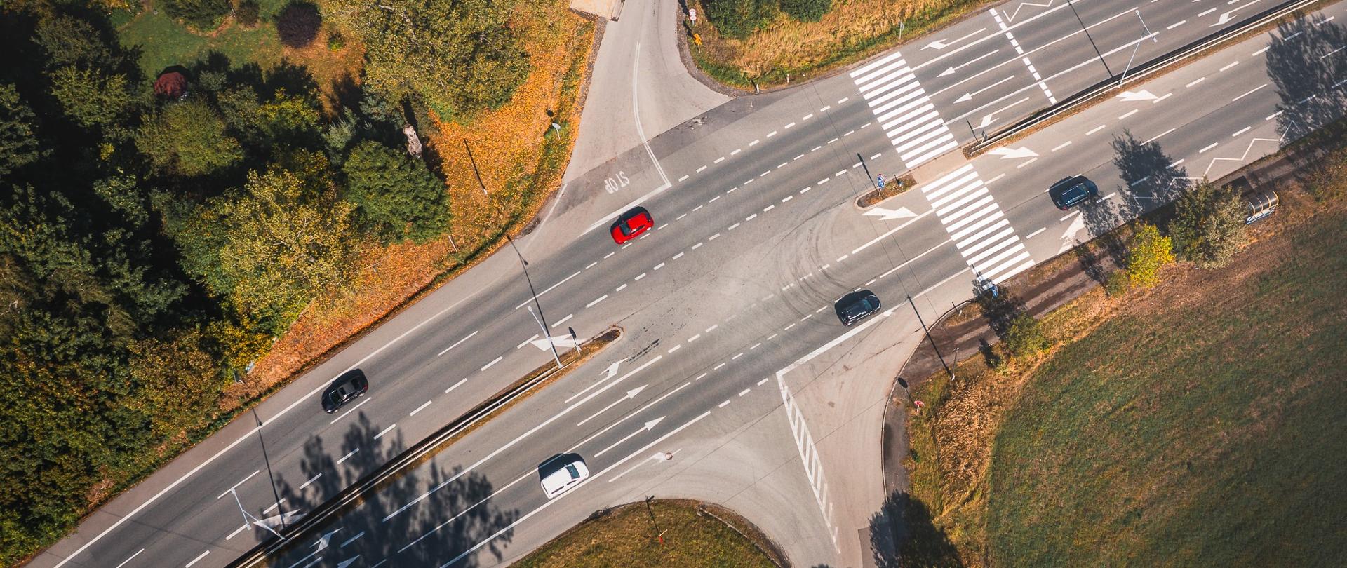 Zdj̨ęcie lotnicze skrzyżowania na drodze DK81, z kilkoma pasami ruchu oraz przejściem dla pieszych. Widoczne samochody poruszające się w obu kierunkach, otoczenie stanowią tereny zielone i zabudowa jednorodzinna.