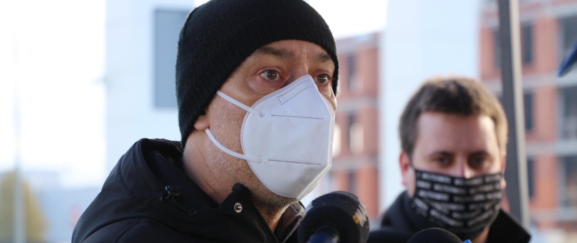 Na zdjęciu wojewoda opolski w maseczce ochronnej, czarnej czapce na głowie, ubrany w czarną kurtkę. Wojewoda stoi przed dziennikarzami i udziela odpowiedzi na pytania dziennikarzy, podczas konferencji prasowej. Ręce wojewody skierowane są ku jego klatce piersiowej. W tle pozostałe osoby, biorące udział w konferencji. 