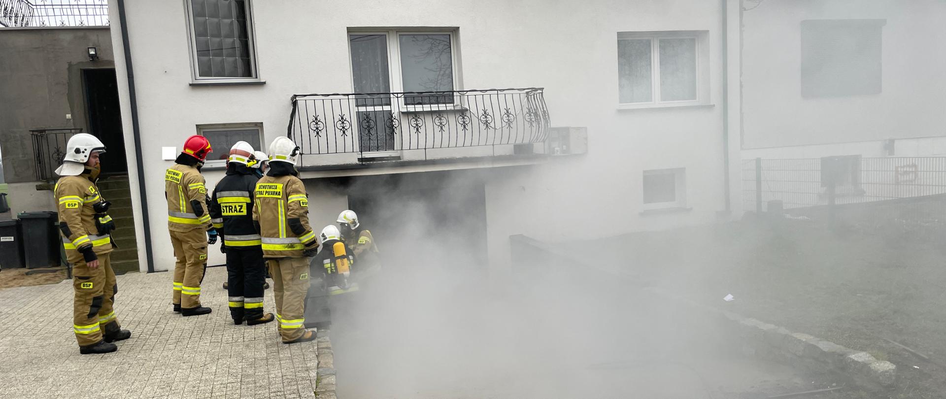 Strażacy stoją przy wejściu do garażu z którego wydobywa się dym