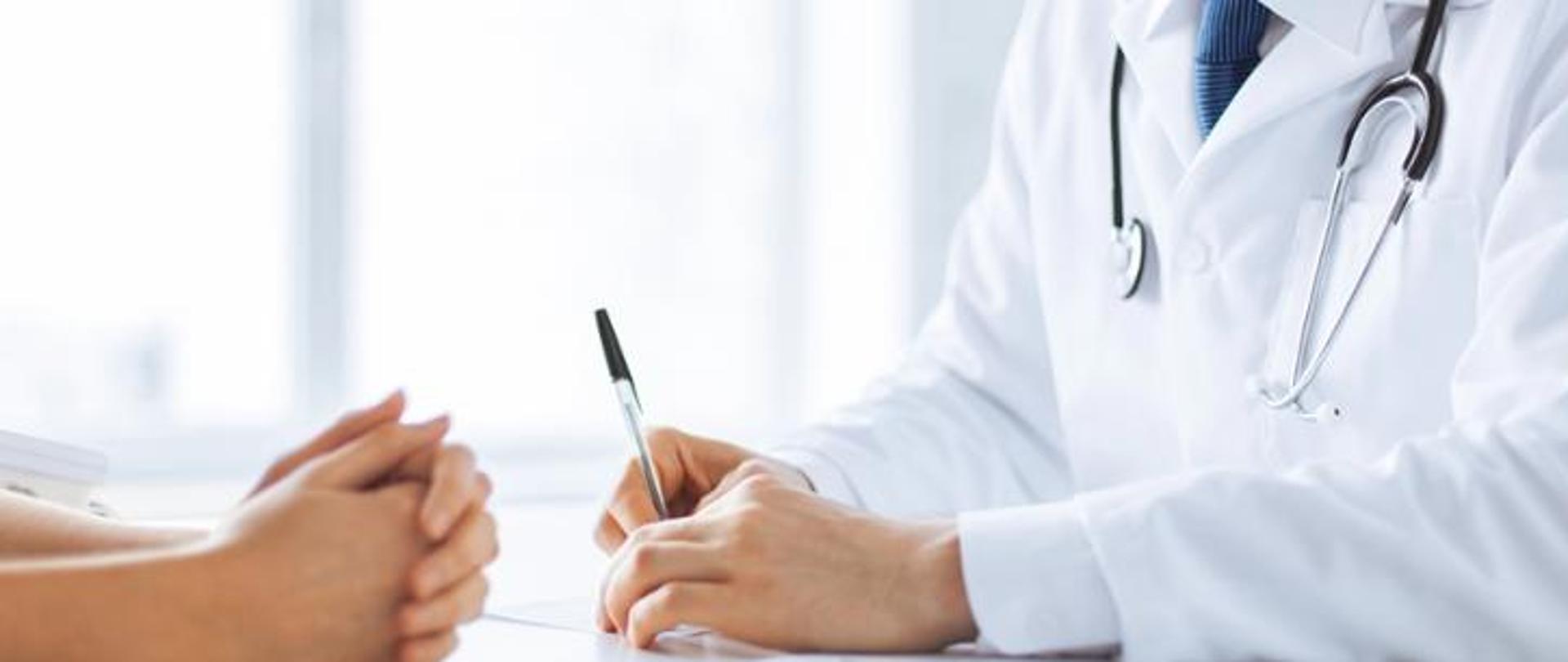 Zdjęcie przedstawia dłonie dwóch osób oparte o biurko. Jedna osoba ubrana w biały kitel widoczna tylko do ramion. Widać stetoskop przewieszony przez szyje i niebieski krawat pod kitlem