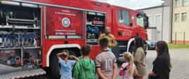 Na zdjęciu grupka dzieci słuchająca strażaka, który opowiada im o sprzęcie znajdującym się na wyposażeniu JRG. Za strażakiem czerwony samochód z naklejką na której znajduje się czujka i napis: Zachowaj bezpieczeństwo, zamontuj czujkę dymu i czadu".