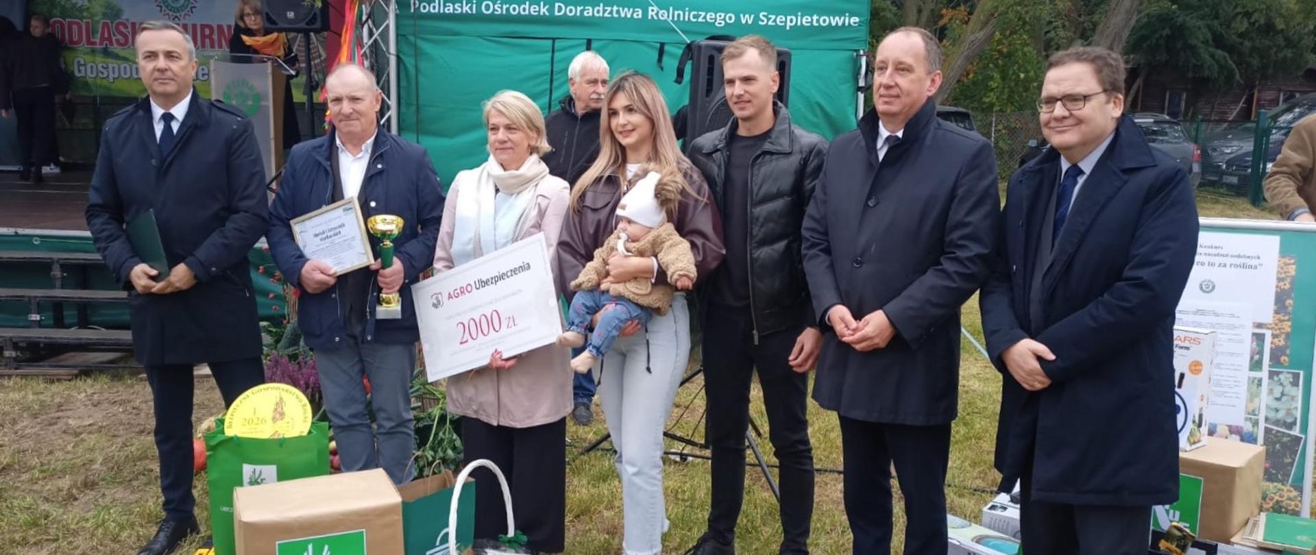 Grupa siedmiu dorosłych osób i małe dziecko pozują do zdjęcia podczas finału etapu wojewódzkiego XXIII Ogólnokrajowego Konkursu „Bezpieczne Gospodarstwo Rolne” w Szepietowie. Wśród uczestników znajdują się zdobywcy pierwszego miejsca – Mariola i Krzysztof Markowscy z miejscowości Brzozowo Antonie. Jeden z laureatów trzyma dyplom i złoty puchar, a stojąca obok kobieta prezentuje bon o wartości 2000 zł. Młoda kobieta trzyma na rękach dziecko. Przed grupą ustawiono nagrody rzeczowe, w tym walizki narzędziowe, torby i upominki od fundatorów. W tle znajduje się scena oraz zielony namiot Podlaskiego Ośrodka Doradztwa Rolniczego w Szepietowie.