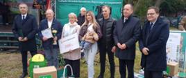 Grupa siedmiu dorosłych osób i małe dziecko pozują do zdjęcia podczas finału etapu wojewódzkiego XXIII Ogólnokrajowego Konkursu „Bezpieczne Gospodarstwo Rolne” w Szepietowie. Wśród uczestników znajdują się zdobywcy pierwszego miejsca – Mariola i Krzysztof Markowscy z miejscowości Brzozowo Antonie. Jeden z laureatów trzyma dyplom i złoty puchar, a stojąca obok kobieta prezentuje bon o wartości 2000 zł. Młoda kobieta trzyma na rękach dziecko. Przed grupą ustawiono nagrody rzeczowe, w tym walizki narzędziowe, torby i upominki od fundatorów. W tle znajduje się scena oraz zielony namiot Podlaskiego Ośrodka Doradztwa Rolniczego w Szepietowie.