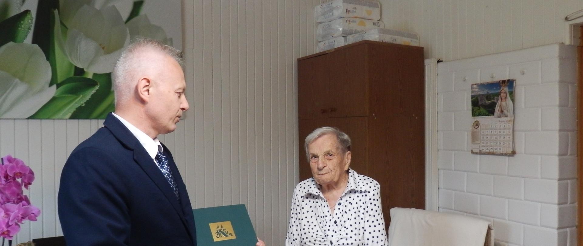 Mężczyzna w garniturze wręczający List gratulacyjny starszej kobiecie stojącej przy stole