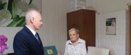 Mężczyzna w garniturze wręczający List gratulacyjny starszej kobiecie stojącej przy stole