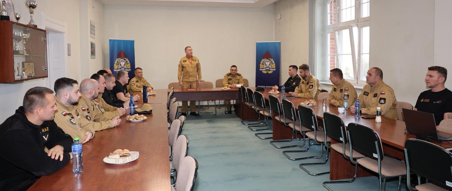 Zdjęcie przedstawia dużą grupę strażaków zgromadzonych w jasnym pokoju konferencyjnym, podczas spotkania narady. W pomieszczeniu znajduje się kilkanaście osób. Większość z nich ubrana jest w mundury w kolorze piaskowym.