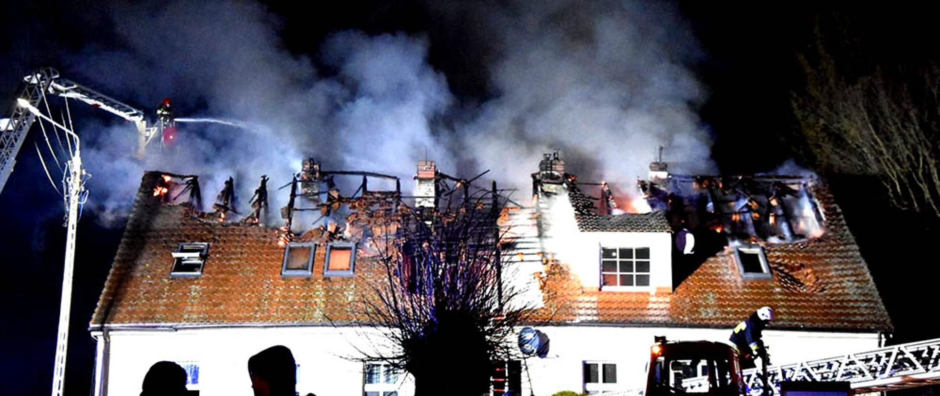 Zdjęcie przedstawia budynek wielorodzinny na którym płonie dach. Straż prowadzi akcję gaśniczą.