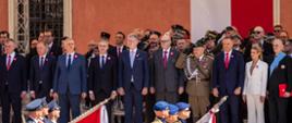 Na zdjęciu uroczystości państwowe z okazji 235. rocznicy uchwalenia Konstytucji 3 Maja. Na zdjęciu trybuna honorowa z najważniejszymi gośćmi. Po prawej stronie zdjęcia w pierwszym rzędzie widać parę prezydencką Karola i Martę Nawrockich oraz Andrzeja Poczobuta. Po lewej stronie zdjęcia minister Tomasz Siemoniak, minister Krzysztof Gawkowski, wiceminister Czesław Mroczek. W tle zdjęcia front Zamku Królewskiego w Warszawie (widać okno po lewej stronie oraz wiszące biało-czerwone barwy narodowe po prawej stronie). Na dole zdjęcia defilada. W pierwszym rzędzie maszeruje trzech żołnierzy w mundurach ze sztandarem. W drugim rzędzie maszeruje trzech policjantów ze sztandarem.