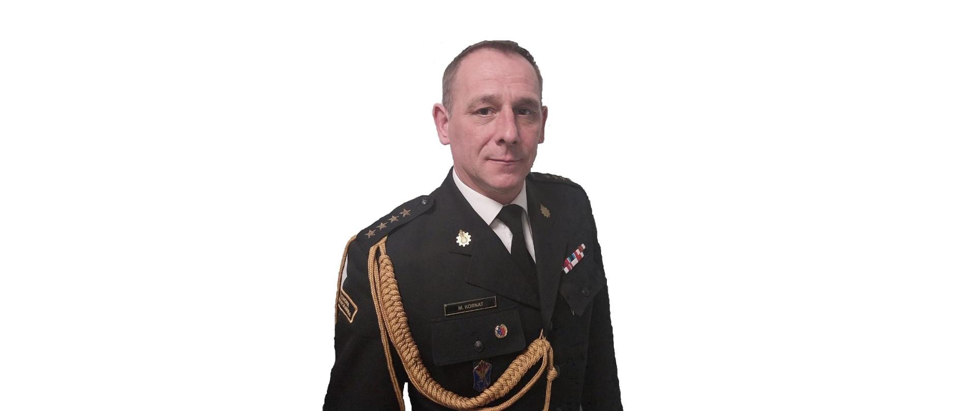 Zdjęcie przedstawia pełniącego obowiązki komendanta powiatowego Państwowej Straży Pożarnej w Łęczycy starszego kapitana Marcina Kornata w czarnym mundurze, na którym znajduje się stopień służbowy oraz odznaczenia strażackie. Zdjęcie portretowe na białym tle.