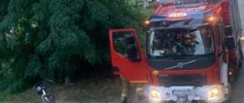 Na zdjęciu znajduje się samochód strażacki z włączonymi światłami błyskowymi, w tle widać inne samochody strażackie oraz drzewa