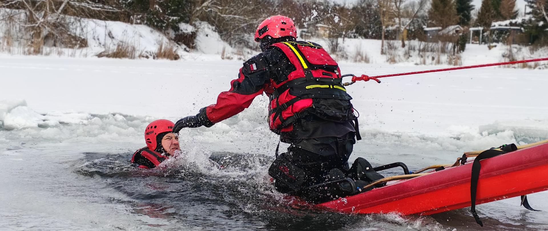 Zdjęcie przedstawia przemyskich strażaków podczas ćwiczenia z zakresu ratownictwa wodno-lodowego , strażak ubabrany w skafander suchy i kask ochronny na saniach lodowych asekurowany linką podejmuje z przerębla drugiego strażaka-pozoranta