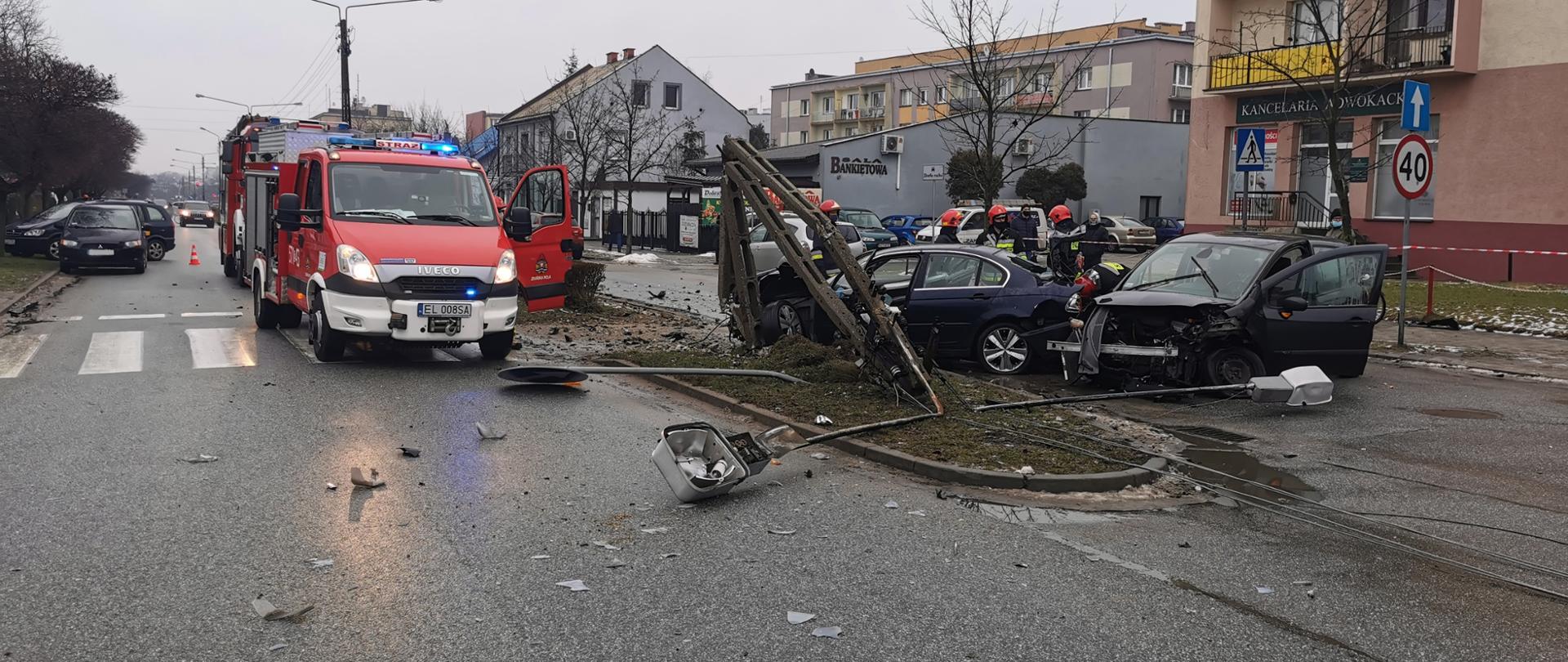 Na zdjęciu widoczni są strażacy zabezpieczający miejsce zderzenia dwóch pojazdów osobowych oraz uszkodzony słup z linią energetyczną. 