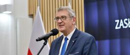W dużej sali stoi wiceminister Murdzek i mówi do mikrofonu na stojaku, za nim pod ścianą polska flaga.