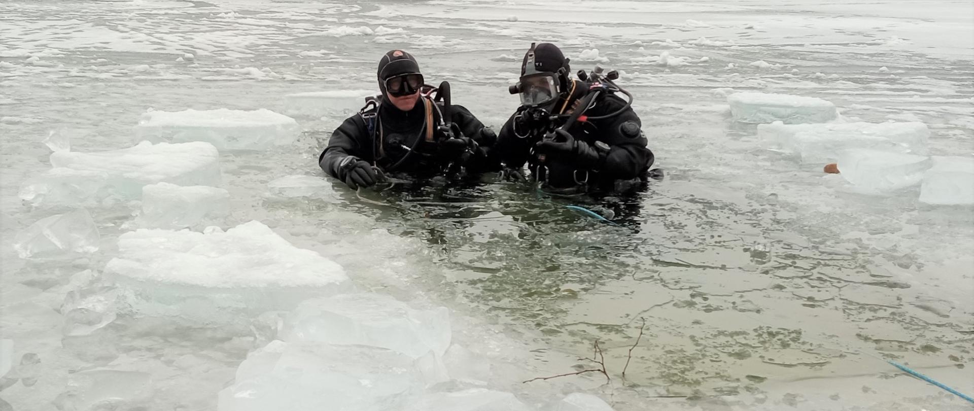Na zdjęciu widzimy dwóch ratowników-nurków w przeręblu. Po lewej stronie instruktor zabezpieczający nurka schodzącego pod lód