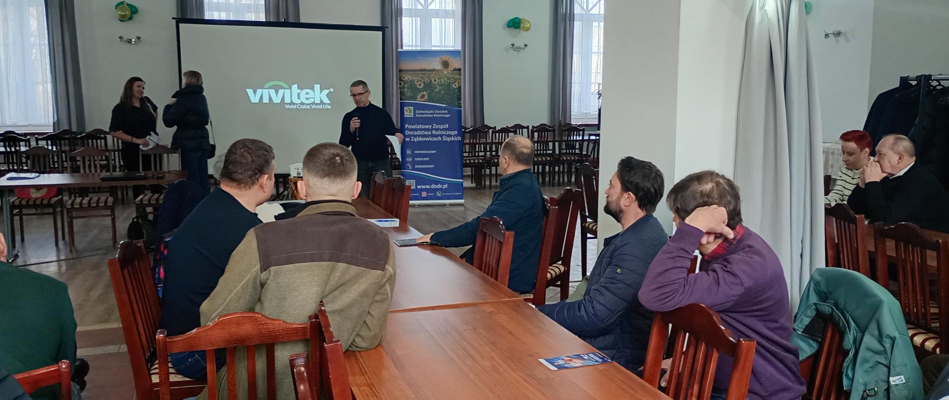 Uczestnicy spotkania siedzą przy stole konferencyjnym a w tle widoczny ekran z prezentacją i wykładowca