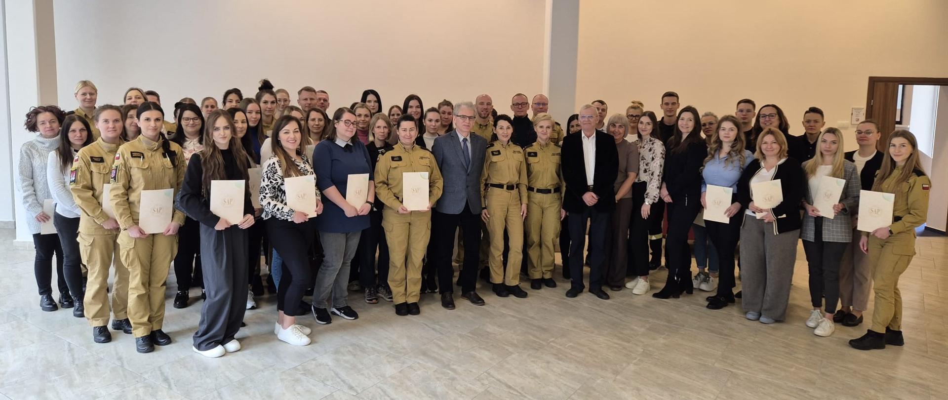 Grupa kilkudziesięciu osób – w tym strażacy PSP w umundurowaniu oraz osoby cywilne – pozuje do wspólnego zdjęcia w sali konferencyjnej. Część uczestników trzyma w rękach dokumenty lub dyplomy.