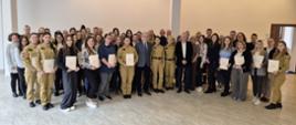 Grupa kilkudziesięciu osób – w tym strażacy PSP w umundurowaniu oraz osoby cywilne – pozuje do wspólnego zdjęcia w sali konferencyjnej. Część uczestników trzyma w rękach dokumenty lub dyplomy.
