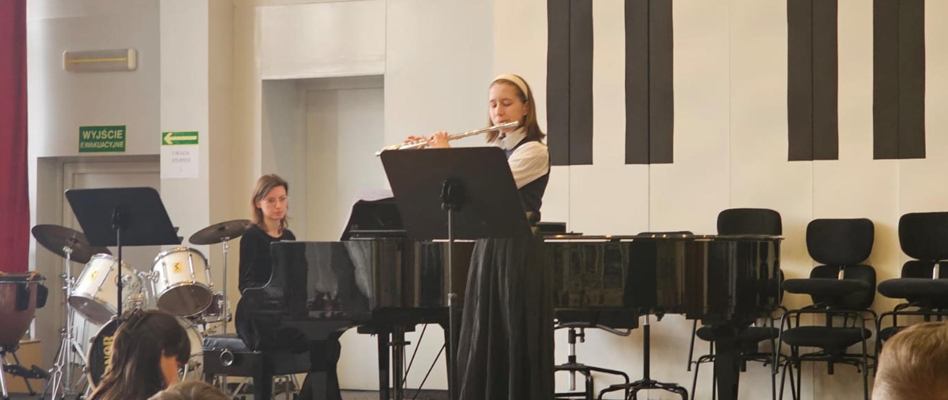 Dziewczynka gra na flecie, kobieta jej akompaniuje na fortepianie.
