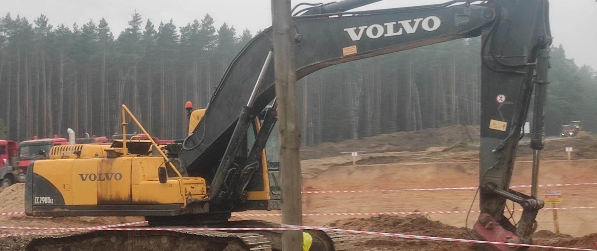 Na zdjęciu koparka Volvo koloru żółtego ramię koparki koloru czarnego. Koparka stoi na piasku z tył samochody oraz las.