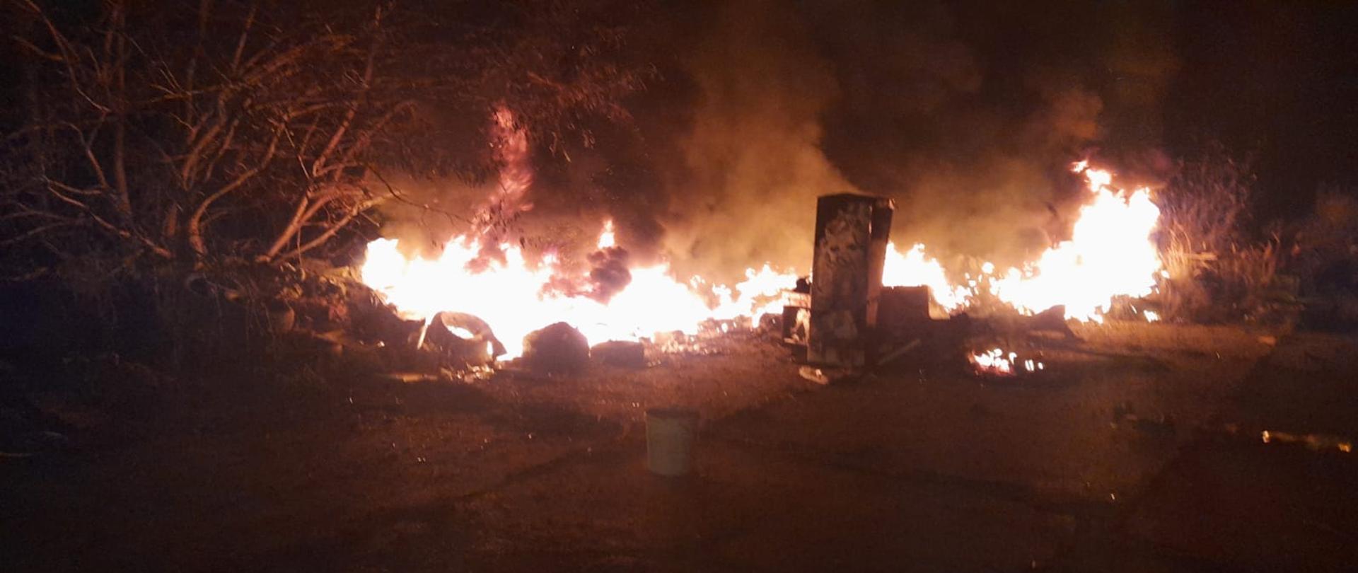 Na zdjęciu widać palące się dzikie wysypisko śmieci na którym znajdują się stare opony oraz śmieci. W pobliżu ognia rosną krzewy. Zdjęcie zostało wykonane po zmroku. 