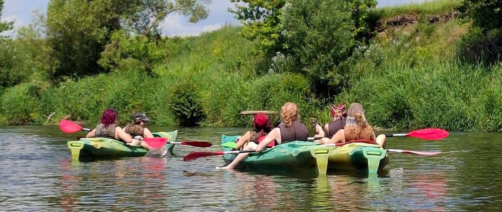 Zdjęcie jest w kompozycji pionowej, na pierwszym planie od dołu zdjęcia do połowy falująca tafla płynącej rzeki, na drugim planie cztery kajaki dryfujące po rzece, osoby siedzące w kajakach mają Wiosła oparte na kolanach. W tle na trzecim planie brzeg rzeki obrośnięty szuwarami i drzewami