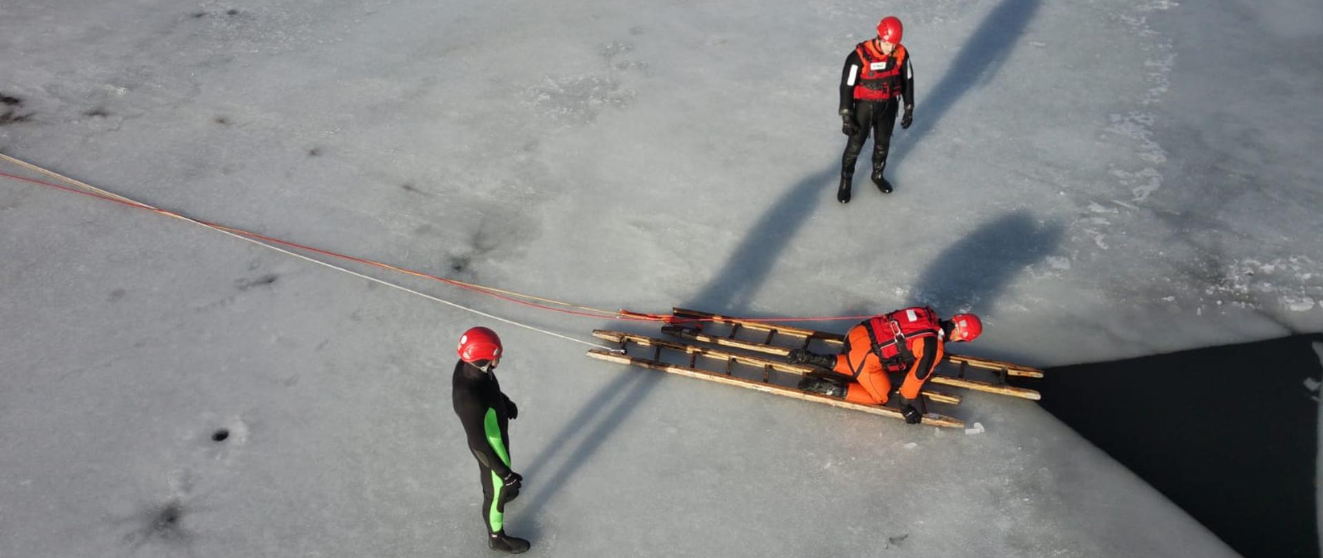 zamarznięte jezioro, na którym znajduje się trzech strażaków ubranych w specjalne kombinezony, jeden z nich klęczy na dwóch przęsłach drabiny