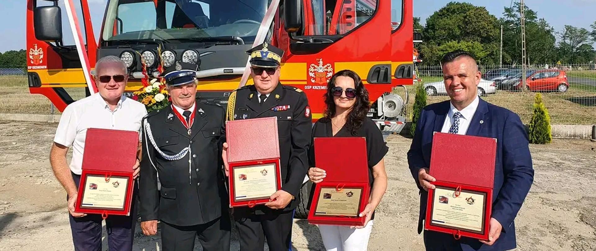 Na tle dużego samochodu strażackiego stoi 5 osób w środku stoi łódzki komendant wojewódzki , po jego lewej stronie pani z rady gminy Dalików obok niej wójt gminy Dalików. Po prawej stronie komendanta stoi przedstawiciel OSP Krzemieniew oraz Poseł na sejm RP Piotr Polak. Trzymają otwarte czerwone pudełka w których pokazują podziękowania 