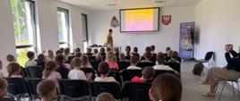 Na pierwszym planie widać dzieci słuchające prelekcji prowadzonej przez znajdującego się na drugim planie strażaka. W tle, na prezentacji widoczne są numery alarmowe 112 oraz 999. 