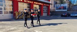 Kolorowa fotografia wykonana w słoneczny dzień na placu Komendy Powiatowej PSP w Brzozowie. Przedstawia poczet flagowy w umundurowaniu wyjściowym z flagą maszerujący w kierunku masztu. W tle strażacy stojący na zmianie służby, jeden z nich oddaje honor.