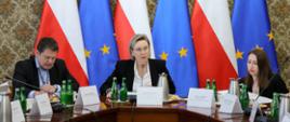 Zdjęcie sytuacyjne. Zbliżenie. Na zdjęciu, przy stole konferencyjnym, widoczni od lewej Pan Łukasz Jachimowicz, Dyrektor Departamentu Społeczeństwa Obywatelskiego w KPRM, Pani Adriana Porowska, Sekretarz Stanu w Kancelarii Prezesa Rady Ministrów, Wiceprzewodnicząca Komitetu do spraw Pożytku Publicznego i Pani Anna Czaplińska, Zastępca Dyrektora w Departamencie Społeczeństwa Obywatelskiego w KPRM. W tle stoją ustawione w rzędzie flagi Polski i Unii Europejskiej.