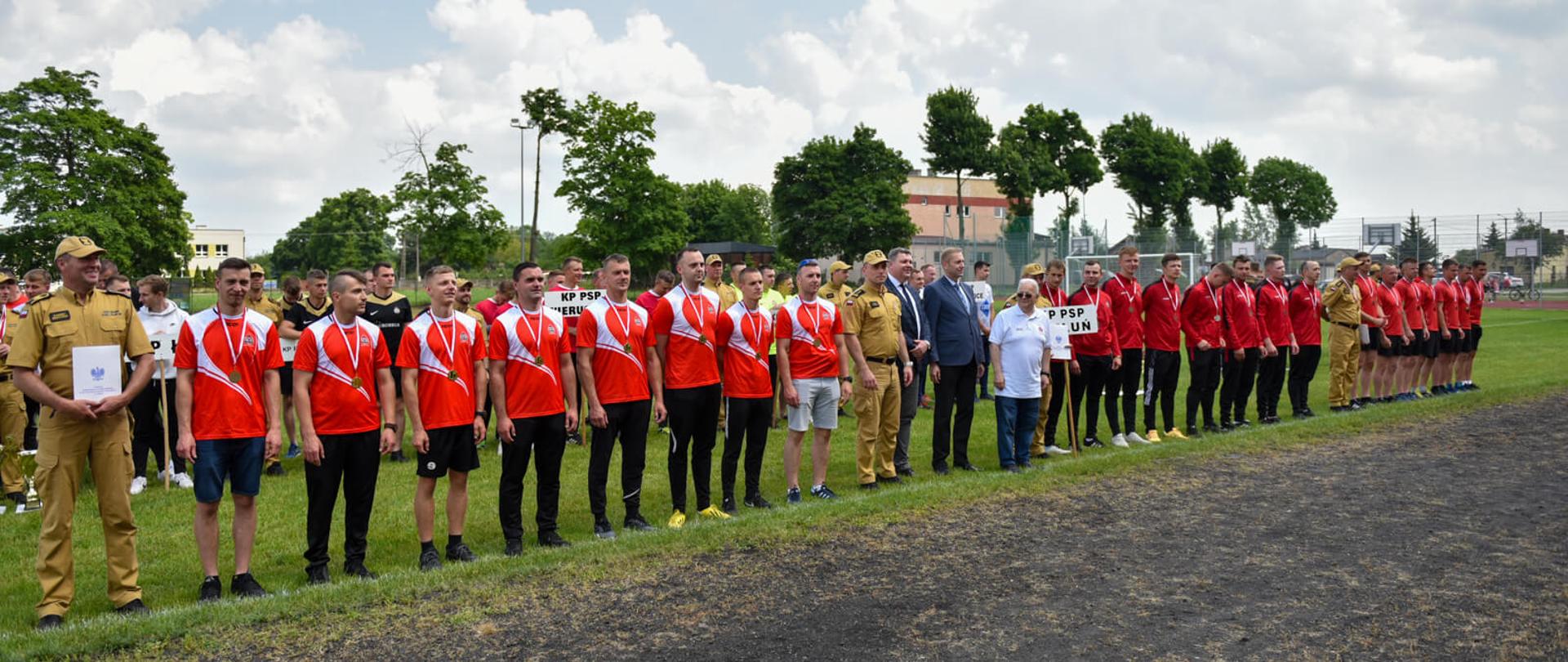 Na zdjęciu widzimy oficjalne zakończenie zawodów. Trzy zwycięskie drużyny pozują do zdjęcia na stadionie piłkarskim. Uczestnicy stoją w rzędzie, są ubrani w stroje sportowe, głównie czerwone koszulki, mają medale na szyjach. Z lewej strony widać mężczyznę w mundurze, który trzyma dyplom. W tle widzimy inne grupy uczestników z tabliczkami wskazującymi przynależność jednostkową. W tle widać drzewa i budynki. 
