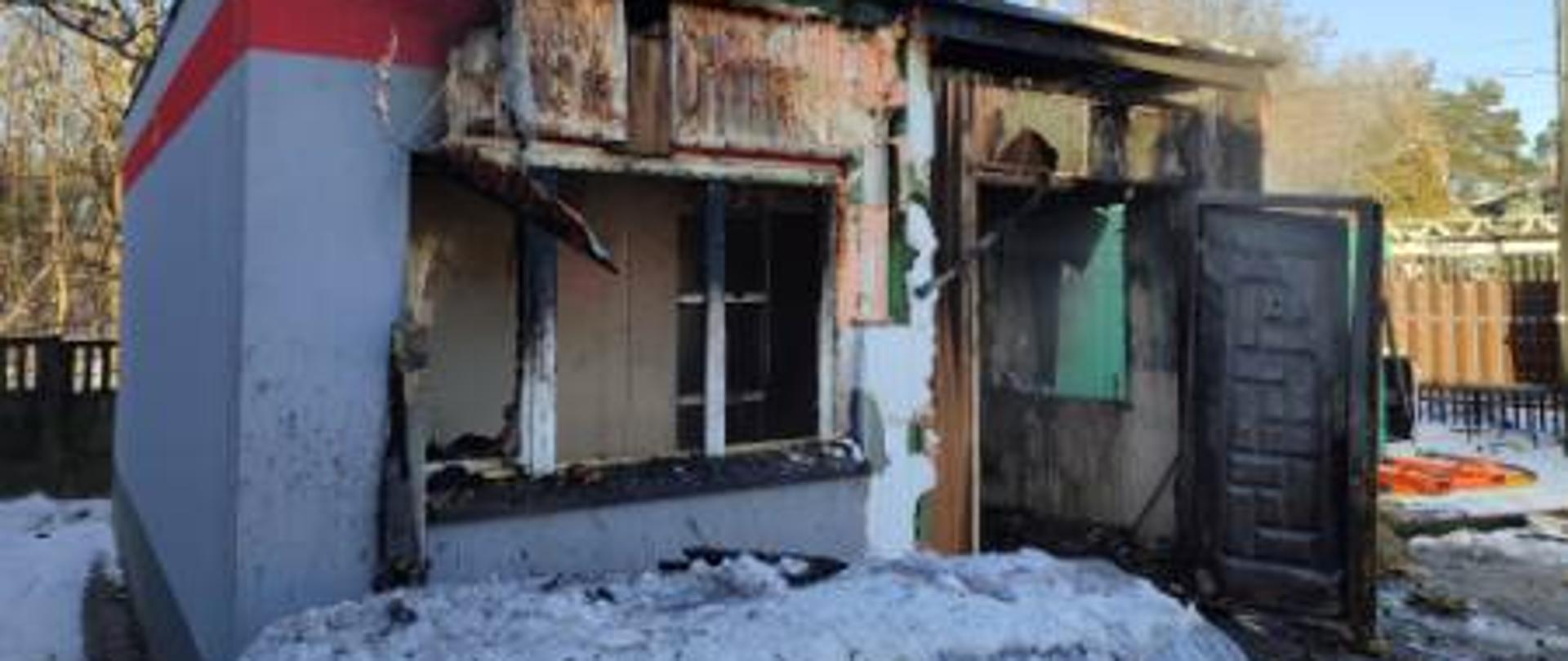 Spalony budynek parterowy, zwęglone drzwi wejściowe oraz część elewacji.
