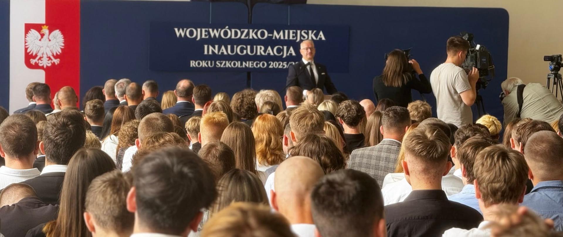 Grupa osób stoi w sali gimnastycznej, w tle jest granatowa ścianka z napisem Wojewódzko-Miejska Inauguracja Roku Szkolnego 2025/2026