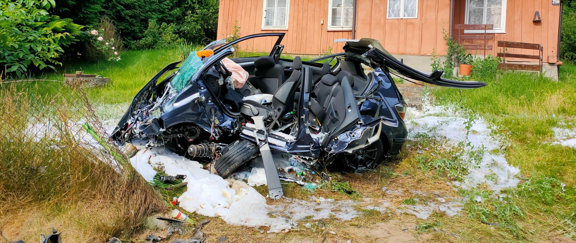 Na zdjęciu widać wrak samochodu osobowego po wypadku, na który podano pianę gaśniczą. Na ziemi znajdują się rozbite częsci pojazdu, a w tle znajduje się drewniany budynek mieszkalny.