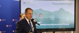 Wiceminister rozwoju pracy i technologii Andrzej Gut-Mostowy stojący przy mównicy z mikrofonem, po jego prawej stronie ekran telewizora z wyświetloną grafiką przedstawiającą dwóch turystów w górach z plecakami i napisem TurystykApp - najlepsza droga do odkrywania usług turystycznych w Polsce.