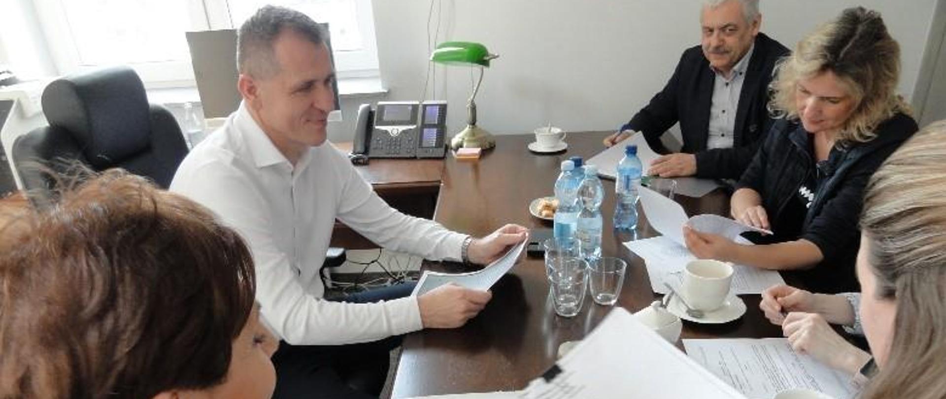 Przy stole siedzi dwóch mężczyzn i trzy kobiety z rozłożonymi na stole dokumentami.