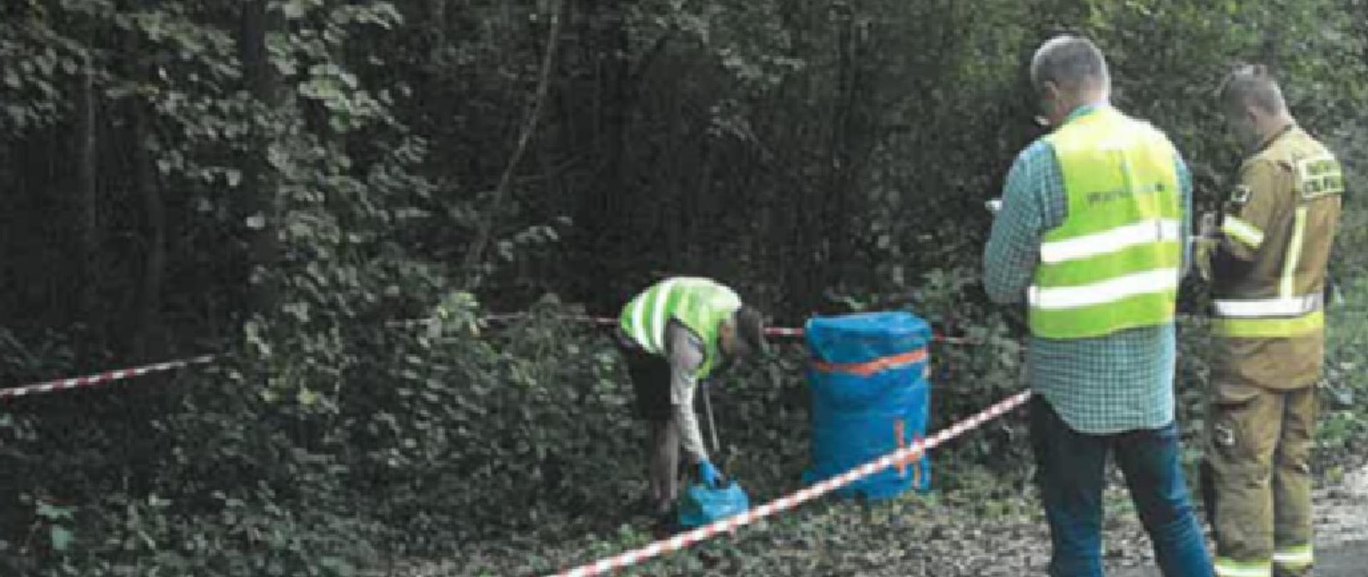 Inspektor Wojewódzkiego Inspektoratu Ochrony Środowiska w Warszawie oraz funkcjonariusz Państwowej Straży Pożarnej stoją na krawędzi drogi i spisują protokoły z działań. Przed nimi, w odległości kilku metrów, za rozwieszoną biało-czerwoną taśmą, na poboczu drogi, stoi zawinięta niebieską folią beczka o pojemności ok. 150 litrów. Obok niej pracownik Centralnego Laboratorium Badawczego Głównego Inspektoratu Ochrony Środowiska pobiera próbkę gleby. W tle widać las.