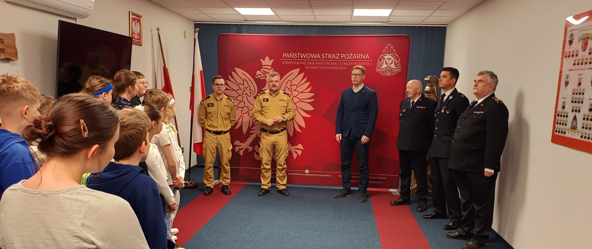 Zdjęcie dokumentuje turniej wiedzy pożarniczej odbywający się w siedzibie Państwowej Straży Pożarnej w Świętochłowicach. W centrum wydarzenia, na tle czerwonej ścianki z godłem, stoi prezydent miasta w asyście funkcjonariuszy służb mundurowych: strażaków w piaskowych ubraniach służbowych oraz druhów OSP w galowych, granatowych mundurach.
Naprzeciwko nich zgromadziła się grupa dzieci i młodzieży biorących udział w konkursie, która z uwagą wysłuchuje oficjalnych wystąpień. Uroczysty charakter spotkania podkreślają obecne w sali symbole narodowe, flaga Polski oraz wyeksponowany w tle historyczny hełm strażacki.
