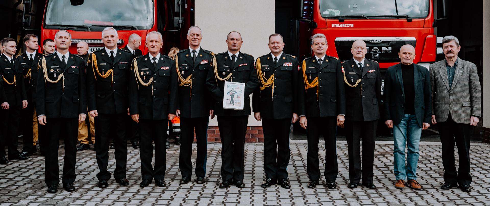 Grupa strażaków stoi do wspólnego zdjęcia przed remizą, pomiędzy dwoma czerwonymi wozami strażackimi. Większość osób ma galowe mundury z odznaczeniami, a jedna z osób w centrum trzyma oprawiony dokument. Zdjęcie ma uroczysty, oficjalny charakter.