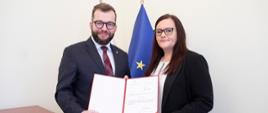 minister Małgorzata Jarosińska-Jedynak i wiceminister Grzegorz Puda trzymają nominację, za nimi flagi Polski i Unii Europejskiej