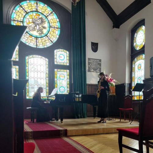 Nastolatka gra na saksofonie, za nią kobieta gra na fortepianie.