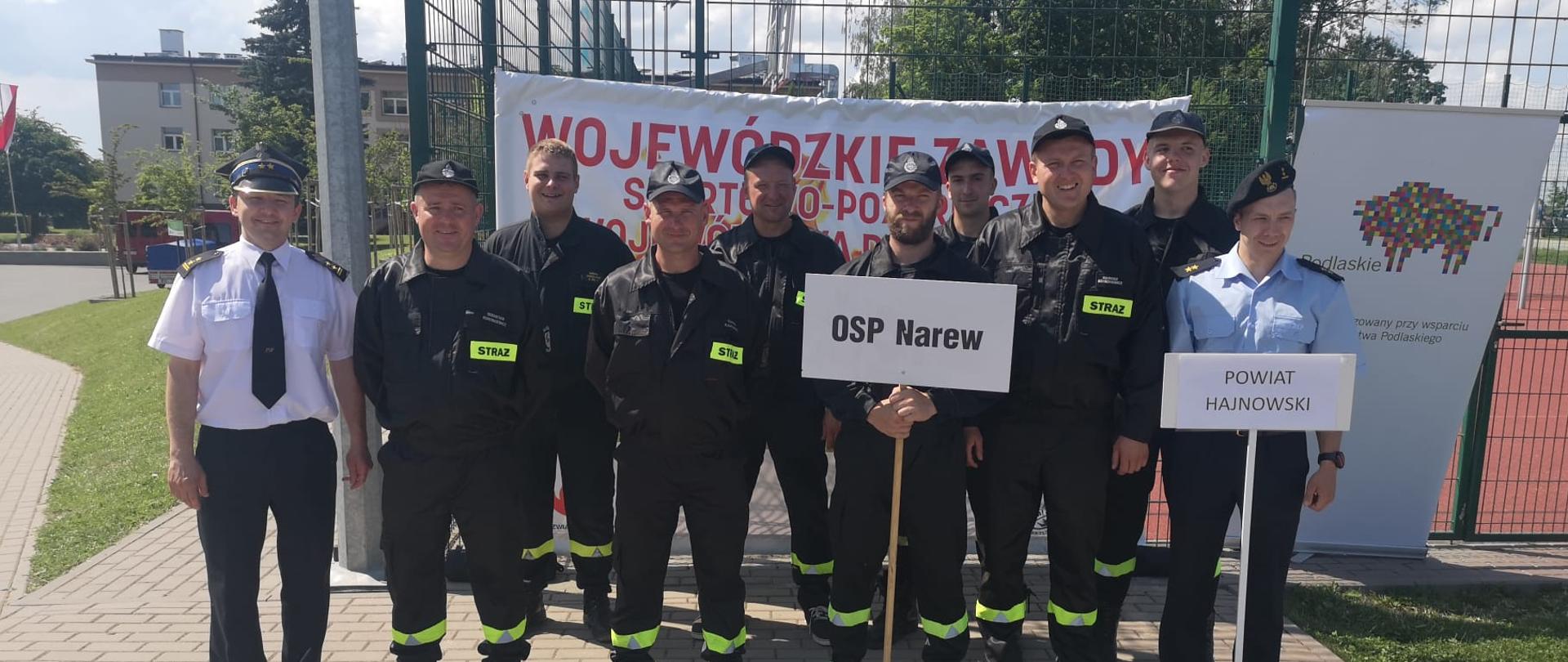 Na tle baneru stoi kilku strażaków w czarnych mundurach po bokach stoją strażacy w niebieskiej i białej koszuli.