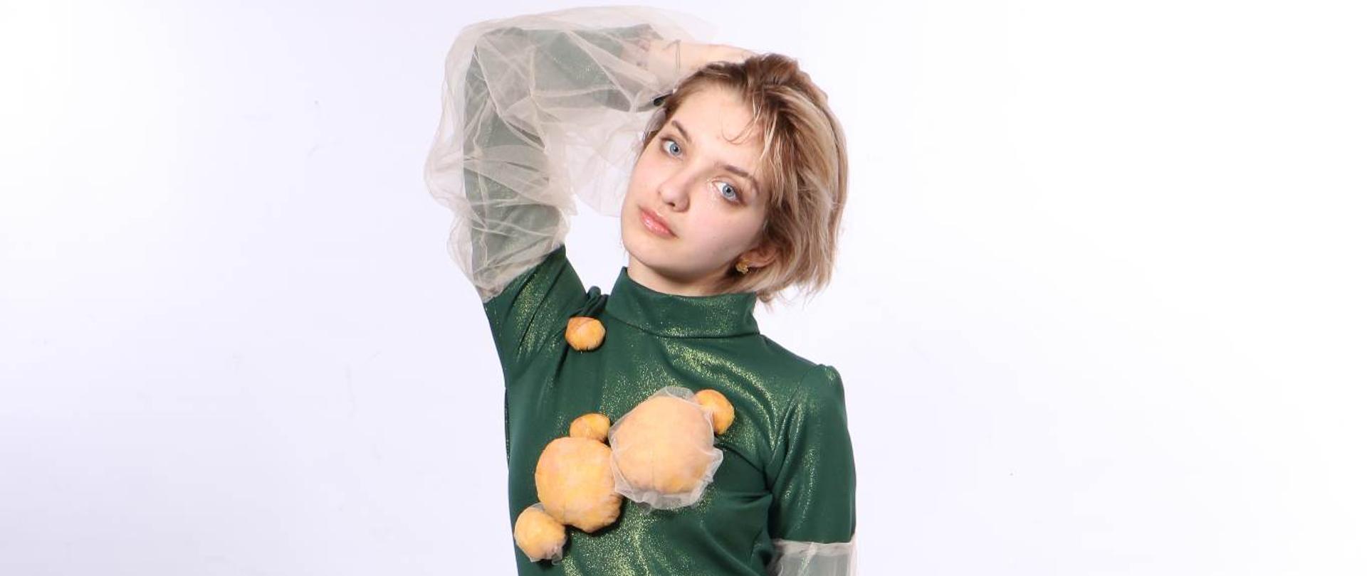 Zdjęcie modelki w zielonym błyszczącym kombinezonie, na wysokości piersi i talii naszyte różnej wielkości pomarańczowe kule z materiału, na rękawach i nogawkach falbany z białego tiulu