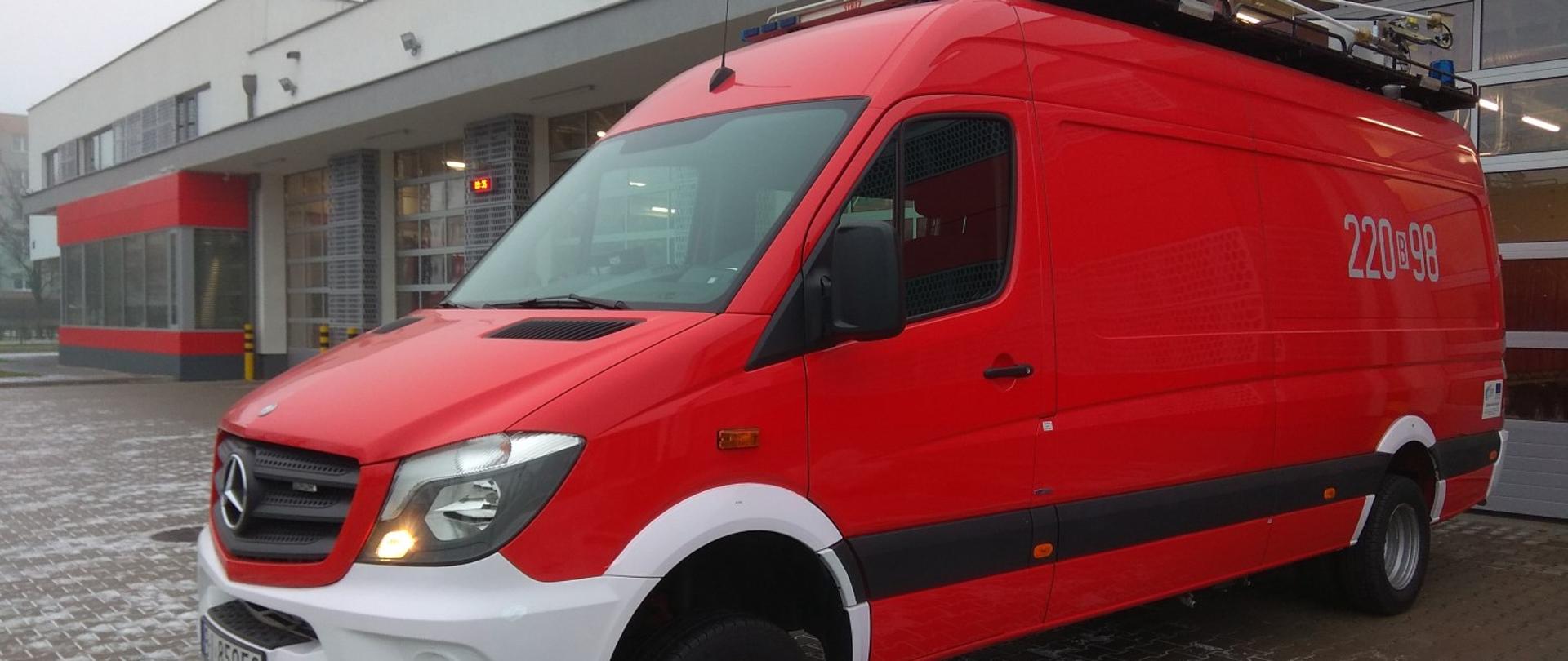 Na pierwszym planie samochód typu bus koloru czerwonego przystosowany do zapewnienia łączności. W tle budynki garażowe.