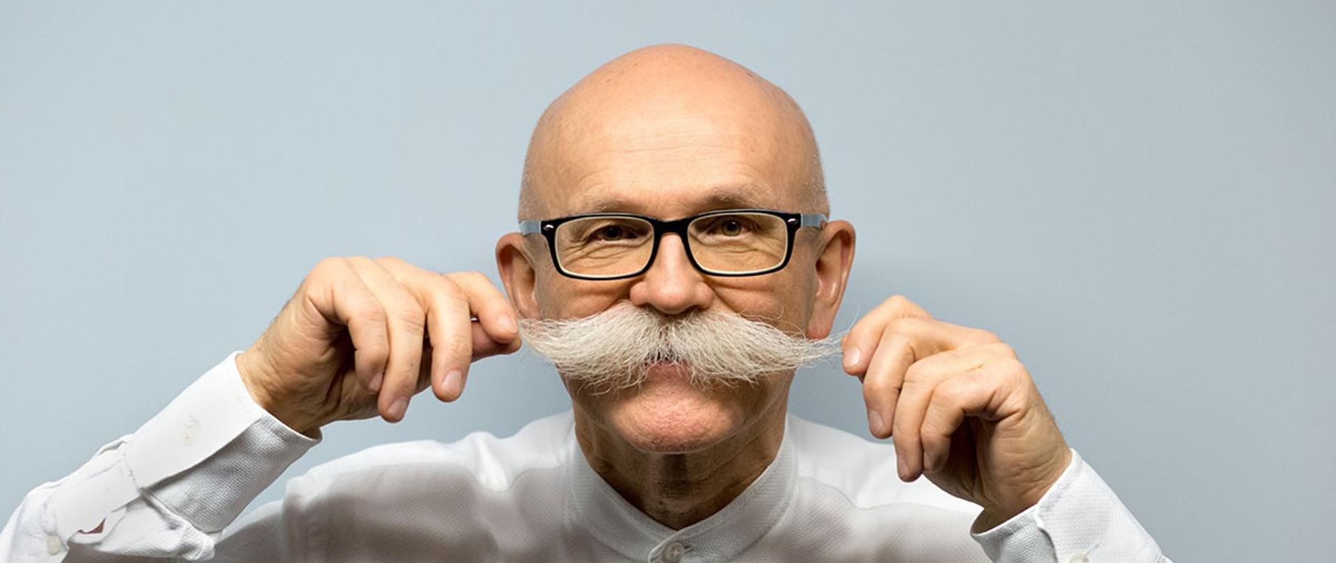 Na zdjęciu widzimy Władysława Grochowskiego, starszego mężczyznę z łysą głową, dużym siwym wąsem oraz okularami w czarnej oprawce. Ma na sobie białą koszulę. Mężczyzna lekko uśmiecha się i jedną ręką podkręca swój wąs. Tło jest jasne i jednolite, bez żadnych dodatkowych elementów.