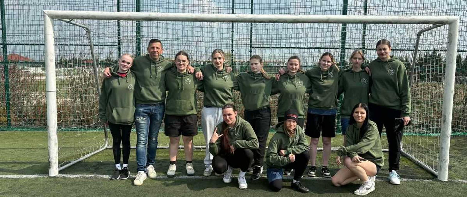 To zdjęcie przedstawia grupę uśmiechniętych osób – prawdopodobnie drużynę sportową – pozujących do wspólnej fotografii na boisku piłkarskim ze sztuczną nawierzchnią.
Oto szczegóły kompozycji:
Grupa osób: W kadrze znajduje się jedenaście osób. Dziewięć z nich stoi w rzędzie, a trzy młode kobiety przykucnęły z przodu. Wśród stojących, po lewej stronie, wyróżnia się mężczyzna, pozostali członkowie grupy to młode kobiety.
Ubiór: Niemal wszyscy mają na sobie identyczne, sportowe bluzy z kapturem w kolorze oliwkowej zieleni. Na lewej piersi bluz widoczne jest okrągłe, jasne logo. Uczestnicy ubrani są w różnorodne spodnie: dresy, krótkie spodenki i legginsy w kolorach czarnym, białym i granatowym.
Bramka: Grupa pozuje bezpośrednio przed białą bramką piłkarską. Siatka bramki jest wyraźnie widoczna, a jej białe słupki i poprzeczka obramowują postacie.
Otoczenie: Akcja dzieje się na zewnątrz. Za bramką znajduje się wysokie ogrodzenie z siatki (piłkochwyt), a za nim widać zarys drzew i budynków w oddali. Niebo jest jasne, z delikatnymi chmurami, co sugeruje pogodny dzień.
Atmosfera: Zdjęcie emanuje pozytywną energią i duchem zespołowym. Osoby stoją blisko siebie, niektóre obejmują się za ramiona, a wszyscy patrzą w stronę aparatu.