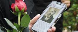 zdjęcie przedstawia zbliżenie na ekran telefonu wyświetlający informacje o zmarłym, widoczny kwiat róży