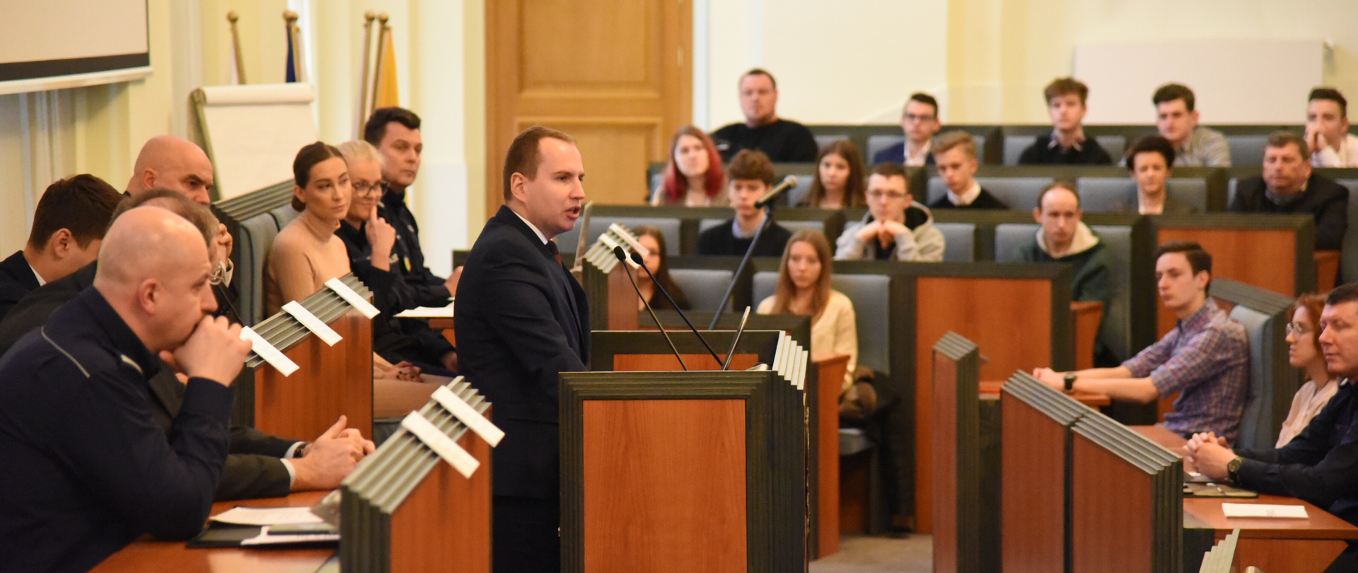 Sala obrad urzędu wojewódzkiego. Minister Adam Andruszkiewicz przemawia przy mównicy. Wokół zgromadzeni są pozostali uczestnicy spotkania. Wśród nich przedstawiciele władz województwa, policji. Przeważają młodzi ludzie