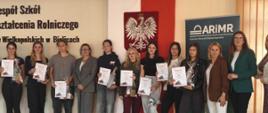 "Zdjęcie przedstawia grupę jedenastu kobiet stojących w rzędzie w sali lekcyjnej lub auli podczas oficjalnego wydarzenia, prawdopodobnie wręczenia nagród.
Tło i otoczenie
• Ściany i napisy: Po lewej stronie, na jasnej ścianie, widnieje czarny napis: „Zespół Szkół Kształcenia Rolniczego [...] Wielkopolskich w Bielicach”. Nad napisem zamontowany jest elektroniczny zegar wskazujący godzinę 15:07.
• Symbole narodowe: W centralnej części tła wisi duża flaga Polski (biało-czerwona), a na niej umieszczone jest godło – Biały Orzeł w koronie na czerwonej tarczy.
• Banner: Po prawej stronie stoi ciemnozielony banner instytucji ARiMR (Agencja Restrukturyzacji i Modernizacji Rolnictwa) z dopiskiem „Kujawsko-Pomorski Oddział Regionalny”.
Osoby na zdjęciu (od lewej do prawej)
Większość dziewcząt po lewej i w środku trzyma w dłoniach pamiątkowe dyplomy oraz drobne, zapakowane w folię upominki.
1. Pierwsza dziewczyna: Młoda kobieta w ciemnym T-shircie, ciemnych spodniach i jasnych butach sportowych. Trzyma dyplom.
2. Druga dziewczyna: Nastolatka z długimi blond włosami, ubrana w czarną bluzę z długim rękawem i ciemne spodnie. Również trzyma dyplom.
3. Trzecia dziewczyna: Młoda osoba w szarym T-shircie i ciemnych spodniach, trzymająca dyplom na wysokości pasa.
4. Czwarta kobieta (starsza): Kobieta w okularach, ubrana w elegancki, szary garnitur (żakiet i spodnie) oraz białą bluzkę. Stoi z rękami splecionymi przed sobą.
5. Piąta dziewczyna: Nastolatka w czarnej bluzie z kapturem i szerokich, szarych jeansach, trzymająca dyplom i upominek.
6. Szósta dziewczyna: Dziewczyna z blond włosami w ciemnozielonej bluzie i bordowych spodniach dresowych, trzyma przed sobą dyplom.
7. Siódma dziewczyna: Młoda kobieta z długimi, ciemnymi włosami, ubrana w czarny T-shirt i jasnoszare spodnie dresowe. Trzyma dyplom w dłoni.
8. Ósma dziewczyna: Dziewczyna w ciemnej bluzie i ciemnych spodniach, trzymająca dyplom.
9. Dziewiąta dziewczyna: Nastolatka w czarnej, skórzanej kurtce (ramonesce) i ciemnych spodniach, trzymająca dyplom i dużą paczkę z nagrodą.
10. Dziesiąta kobieta: Starsza, elegancka kobieta w brązowym żakiecie, białej bluzce i białych spodniach oraz białych butach sportowych.
11. Jedenasta kobieta: Elegancka kobieta w okularach, ubrana w ciemnozieloną (butelkową) marynarkę, czarne spodnie i czarne szpilki.
Po prawej stronie kadru widoczny jest również fragment dwunastej osoby w wielokolorowej, wzorzystej sukience. Cała grupa stoi na jasnej, drewnianej podłodze z paneli i patrzy w stronę obiektywu, większość osób delikatnie się uśmiecha."