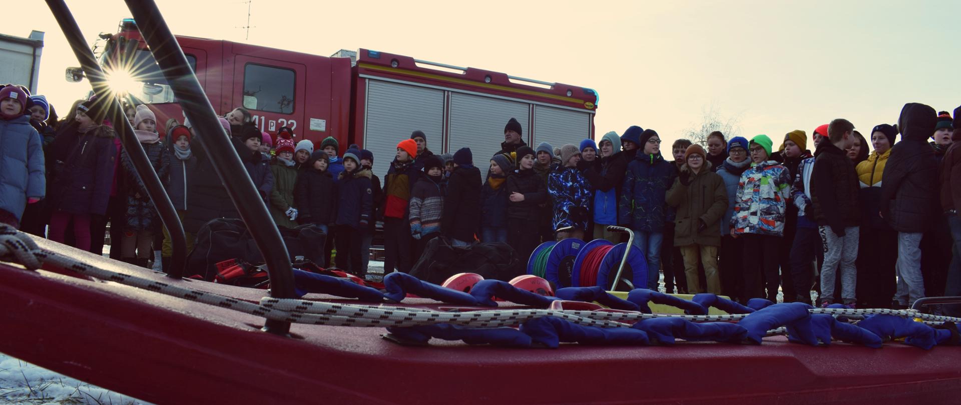 Zdjęcie wykonane na zewnątrz. Wokół dużo śniegu, jest mroźna pogoda. Zdjęcie przedstawia strażaków oraz uczestników akcji „BEZPIECZNE FERIE 2024”